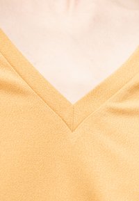 Κοντινό πλάνο ενός κίτρινου πλεκτού τοπ με V-λαιμόκοψη που δείχνει την υφή και το ντεκολτέ σε φόντο ανοιχτού δέρματος.