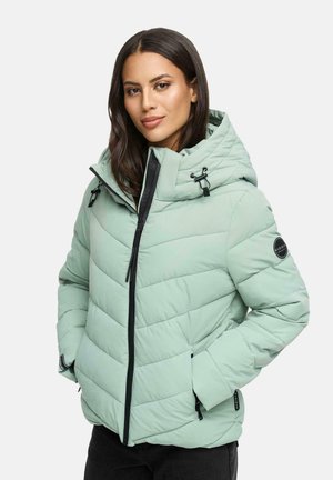 Woman wearing a light green quilted puffer jacket with hood, black zipper, and hands in pockets, standing against a plain background.