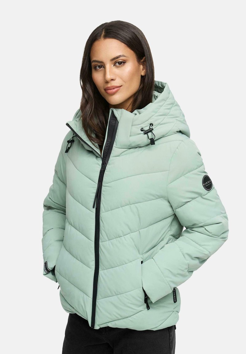 Woman wearing a light green quilted puffer jacket with hood, black zipper, and hands in pockets, standing against a plain background.