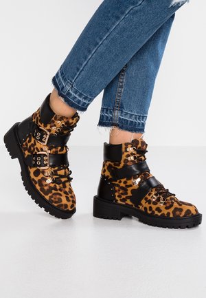 Knöchelhohe Stiefel mit Leopardenmuster, schwarzen Riemen, goldenen Ösen und robuster Sohle, getragen zu Jeans mit ausgefranstem Saum.