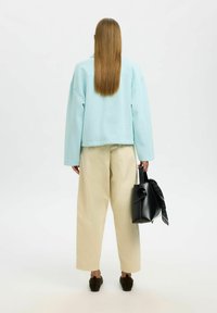 Hellblaues, übergroßes Sweatshirt, cremefarbene Hose mit weitem Bein, braune Schuhe; hält eine schwarze Tasche mit einem auf einer Seite gebundenen gemusterten Schal.