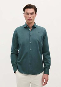 Cămașă butonată de culoare verde închis, cu mâneci lungi, textură moale și tiv rotunjit. Prezintă un logo subtil pe piept și nasturi asortați.