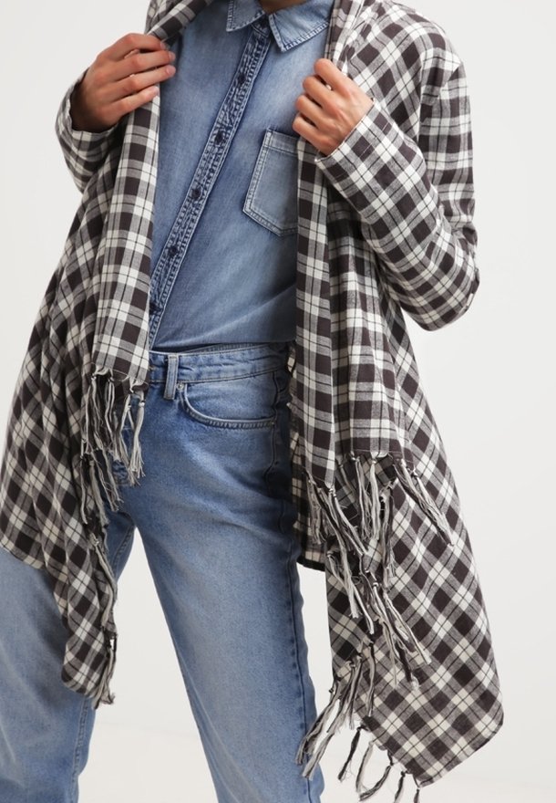 Personne portant une chemise en jean bleue et un jean, tenant ouverte une étole ou une écharpe à franges à carreaux noirs et blancs.