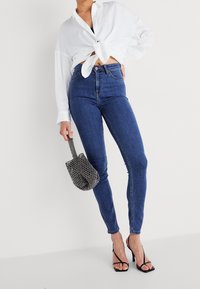 Jeans ajustados de talle alto en un azul oscuro con una textura suave, combinados con una camisa blanca atada y un bolso negro con cuentas. Sandalias de tacón alto negras.