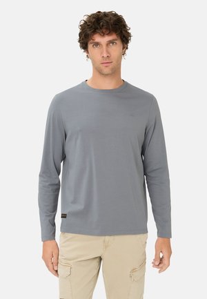 Man met krullend haar, gekleed in een grijs shirt met lange mouwen en beige cargobroek, staand tegen een effen lichte achtergrond.