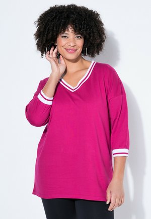 Smilende kvinde med krøllet hår iført creoløreringe og en lyserød langærmet top med hvide kantdetaljer ved halsudskæring og manchetter.