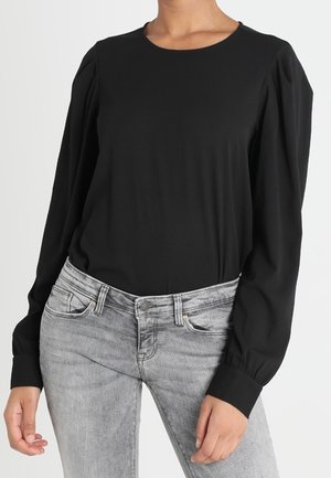 Vrouw draagt een zwart blouse met lange mouwen en opgeblazen schouders, gecombineerd met grijze high-waisted jeans, staand tegen een effen witte achtergrond.