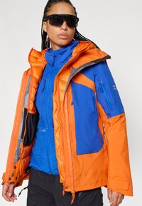 Ένα πολύχρωμο μπουφάν με πορτοκαλί και μπλε υλικά, με καπιτονέ υφή, ρυθμιζόμενη κουκούλα και πολλές φερμουάρ τσέπες για αποθήκευση.
