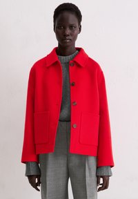 Rode jas van een wolmix met een kraag, knoopsluiting en voorzakken; gedragen over een grijze trui en grijze geruite broek.