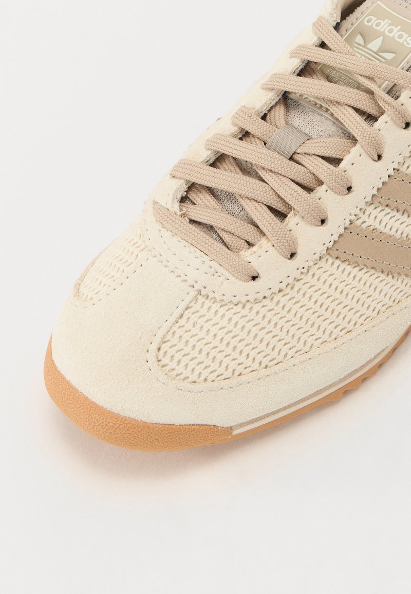 Sneaker beige avec tige en daim et en maille, semelle extérieure texturée et lacets beige. Présente trois rayures marron et une étiquette logo sur la languette.