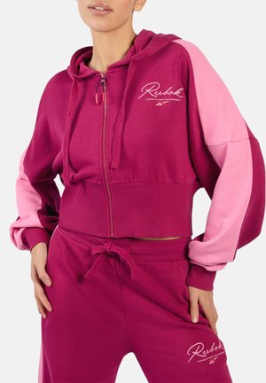 Frau trägt einen zweifarbigen Reebok-Hoodie in Rosa und Fuchsia sowie passende Jogginghosen mit Logotext und Kordelzug an der Taille, die Hände auf den Hüften ruhend.