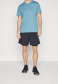 Muž v světle modrém tričku Nike, černých vrstvených šortkách a černých sportovních botách stojí před jednobarevným bílým pozadím.