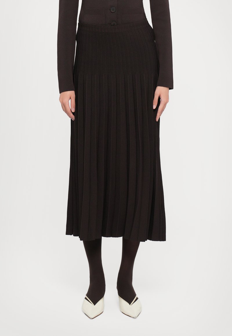 Black pleated midi skirt crafted from knit fabric, featuring a ribbed waistband and paired with dark tights and pointed white shoes.