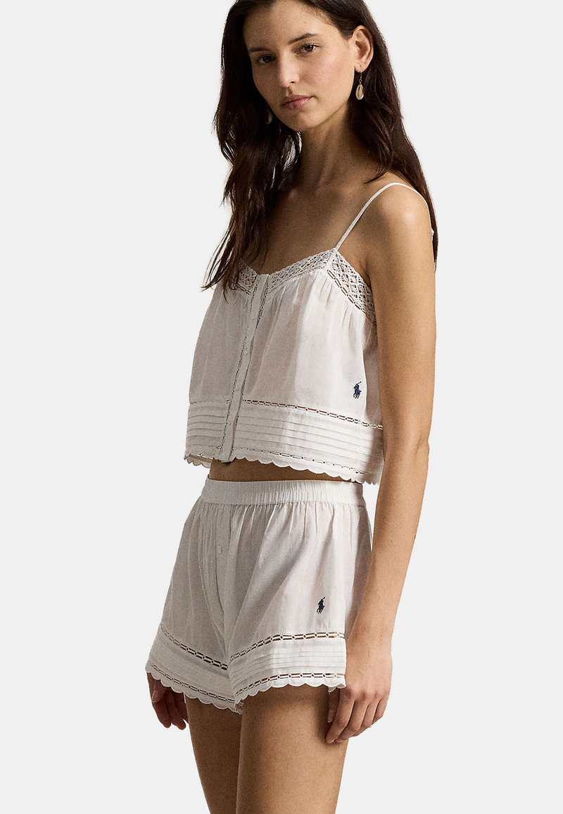 Woman wearing white sleeveless top and matching shorts with lace trim and small embroidered logo on a light background.