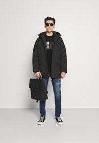 Svart jakke med fyll og glidelås, svart T-skjorte, blå slitte jeans, svarte joggesko, og en svart ryggsekk med spenne-lukkinger.