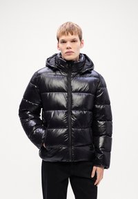 Zwarte pufferjack met een glanzende afwerking, ritssluiting aan de voorkant en capuchon. Heeft een gequilt ontwerp met een puffy textuur en zijzakken.