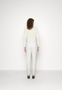 Witte langarmtop met textuurdetails, gecombineerd met aangesloten witte broek. Model draagt zwarte blokhak schoenen. Effen witte achtergrond.