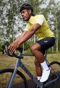 Radfahrer auf einem grauen Canyon-Rad, der ein gelbes Kurzarmtrikot, marineblaue Shorts, weiße Socken und weiße Schuhe trägt, unterwegs auf einem von Bäumen gesäumten Weg.
