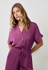 Camicia corta a maniche corte di colore viola con chiusura a bottoni e colletto, caratterizzata da una texture liscia e una vestibilità rilassata, abbinata a pantaloni coordinati.