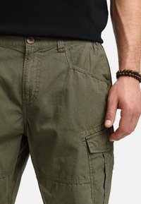 Pantalons cargo vert olive avec fermeture à boutons, poches plaquées et détails de couture visibles. Le tissu a une apparence texturée.