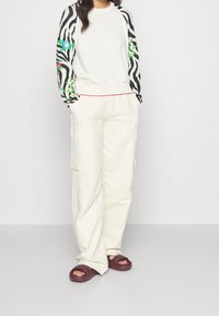 Sweat-shirt blanc avec manches longues à motif noir et floral, associé à un pantalon ample crème et des chaussures maroon à enfiler.