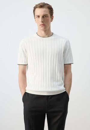 Joven con pelo corto que lleva una camiseta blanca de manga corta acanalada y pantalones negros, de pie con las manos en los bolsillos contra un fondo liso.