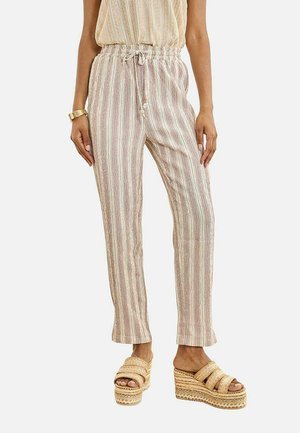 Mujer con pantalones de cordón beige y blanco a rayas y sandalias de plataforma tejidas, de pie con los brazos relajados a los lados.