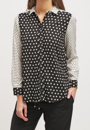 Zwart-witte blouse met aan de ene kant een bloemenpatroon en aan de andere kant stippen; lange mouwen, knoopsluiting aan de voorkant en een kraag.