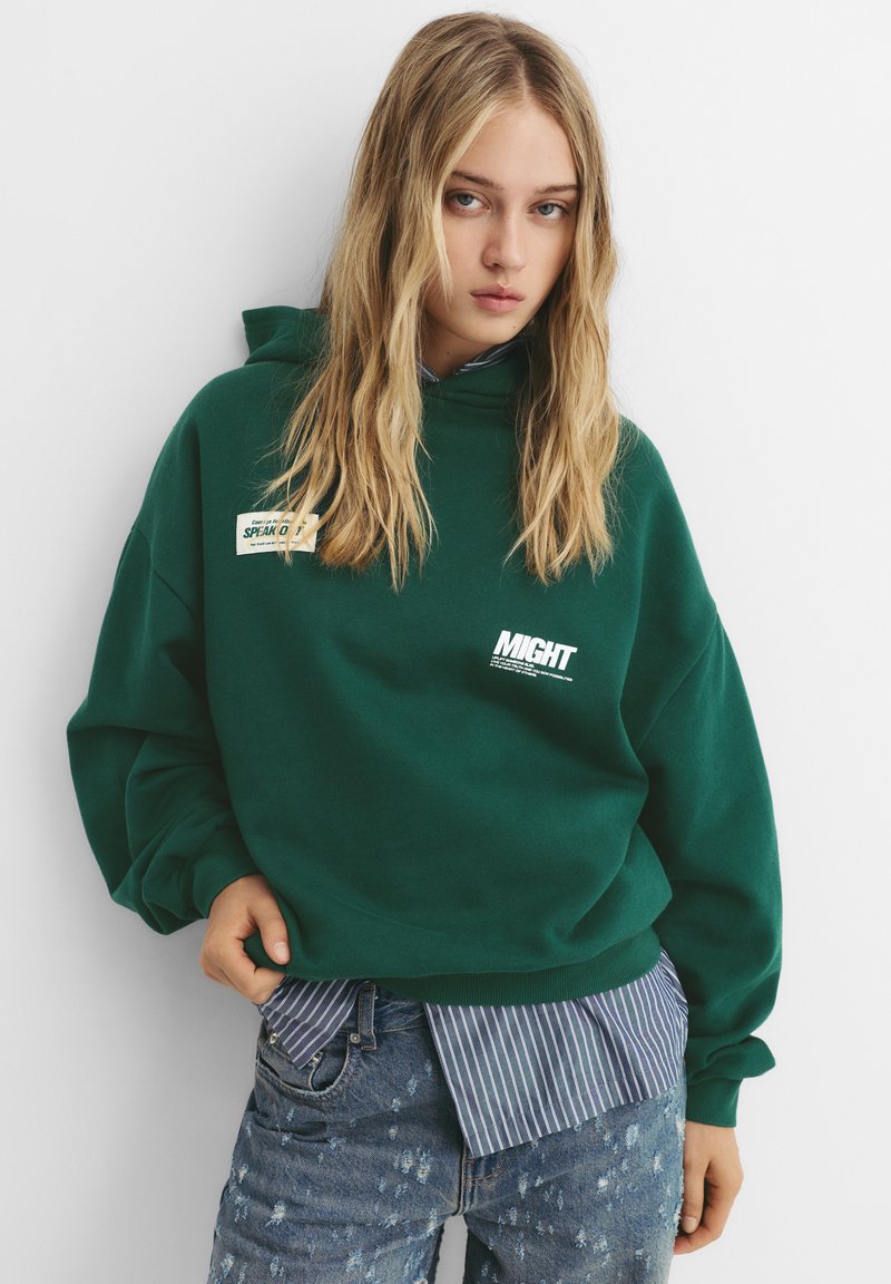 Zielona bluza z kapturem, z naszywkami z przodu; luźny krój. Połączona z niebieską koszulą w paski oraz dżinsami w stylu distressed z lekko zwężającą się nogawką.