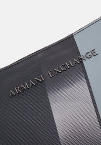 Crna i siva teksturirana torbica s patentnim zatvaračem. Sadrži utisnuto slovo "ARMANI EXCHANGE", s glatkom, sjajnom trakicom kao naglaskom.