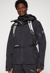 Casaco impermeável preto com colarinho alto, fecho frontal, punhos ajustáveis e vários bolsos. Possui um arnês e tiras refletoras.