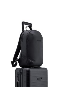 Sac à dos noir avec une finition lisse et mate et des bretelles réglables, placé sur une valise rigide noire avec des rainures horizontales.