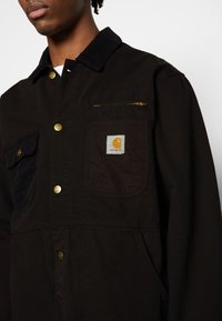 Homme portant une veste Carhartt noire avec des boutons dorés, une poche poitrine zippée et un écusson logo sur la poche gauche, sur fond uni.