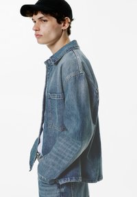 Leren spijkerjack in lichtblauw met een relaxte pasvorm, knoopsluiting aan de voorkant, een enkele borstzak en gestikte details, gedragen over een wit shirt.