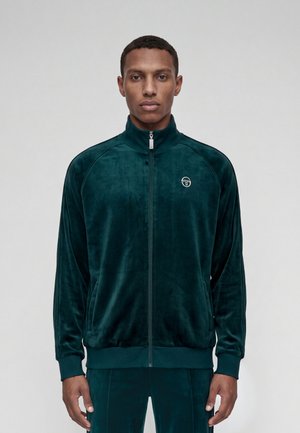 Jeune homme aux cheveux courts porte une veste zippée en velours vert foncé et un pantalon assorti, debout devant un fond clair uni.