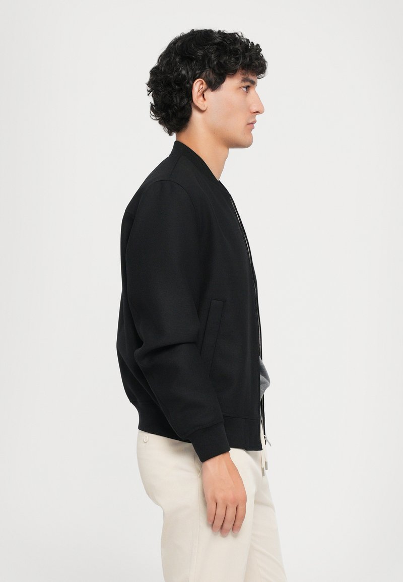 Zwarte bomberjack van textiel, met een rits aan de voorkant, zijzakken en geribbelde manchetten. G gedragen met lichtgekleurde broek.