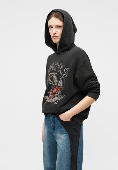 Γυναίκα που φοράει μαύρο φούτερ με κουκούλα με γραφικό απεικονισμένο ένα ράπισμα πάνθηρα και κόκκινα τριαντάφυλλα, συνδυασμένο με τζιν δύο αποχρώσεων σε μπλε και μαύρο.