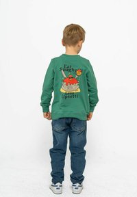 Grön tröja med en färgglad spaghetti-grafik och texten "Ät spaghetti, mindre uppsatt". Bärs med blå jeans och vita sneakers.