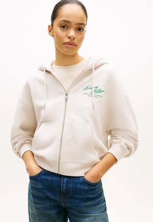 Junge Frau trägt eine hellbeige Zip-Up-Hoodie mit grünem Tommy Hilfiger Logo und blauer Jeans, Hände in den Taschen, neutraler Hintergrund.