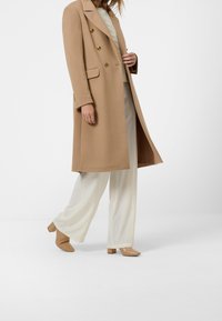 Abrigo beige de mezcla de lana con doble botonadura y botones en tono dorado, combinado con pantalones de pierna ancha en color crema y zapatos beige con tacón ancho.