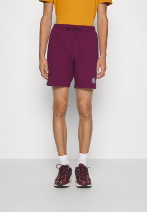 Hombre con pantalones cortos morados con cordón, camiseta amarillo mostaza, calcetines blancos y zapatillas de correr burdeos, de pie contra un fondo liso.