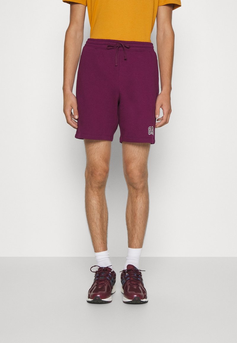 Homme portant un short violet avec cordon, une chemise jaune moutarde, des chaussettes blanches et des chaussures de course bordeaux, debout devant un fond uni.