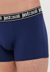 Jūras zēnu bokseršortus ar melnbaltu "Justcavalli" logotipa jostu, pielāgotu dizainu un gludu auduma tekstūru.