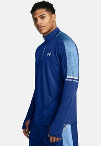 Pitkähihainen urheilupaita tummansinisessä värissä, jossa on puolivetoketju kauluksessa. Paitassa on vaaleansiniset yksityiskohdat olkapäällä ja Under Armour -brändäys hihassa.