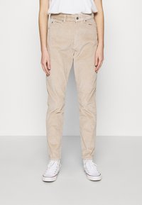 Osoba v béžových manšestrových kalhotách, bílých teniskách a bílé košili zasunuté do kalhot, stojící na neutrálním světlém pozadí.