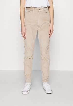 Persona con pantalones de pana beige, zapatillas blancas y una camisa blanca metida por dentro, de pie contra un fondo claro y liso.