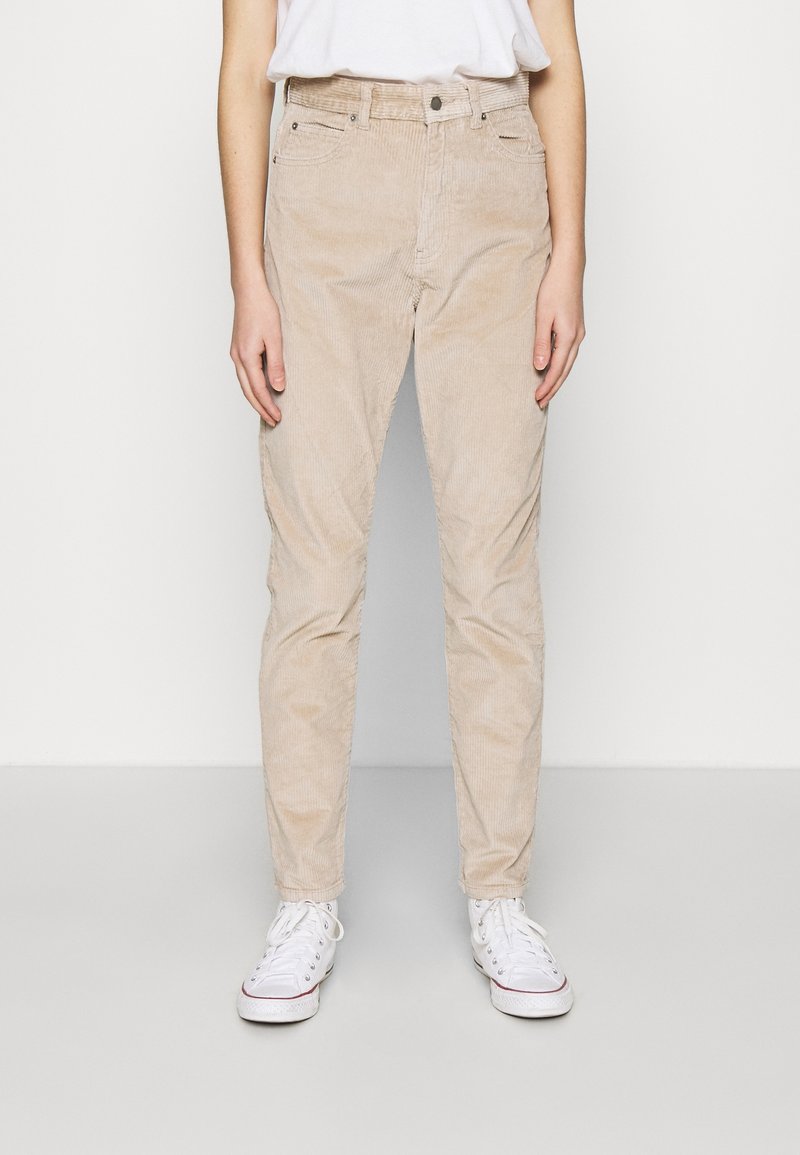 Osoba v béžových manšestrových kalhotách, bílých teniskách a bílé košili zasunuté do kalhot, stojící na neutrálním světlém pozadí.