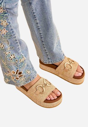Pies con sandalias de plataforma beige tejidas con anillos dorados combinadas con vaqueros azules con bordado floral en el dobladillo.