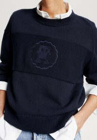 Person, der einen marineblauen Strickpullover mit einem erhabenen runden Logo über einem weißen Hemd trägt, die Hände in den Taschen einer hellblauen Jeans.