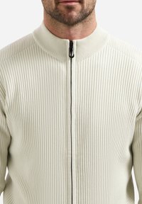 Man in een crèmekleurige geribbelde zip-up trui met een zwarte ritssluiting, weergegeven van schouders tot kin tegen een witte achtergrond.